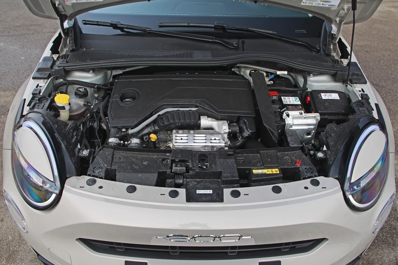 silnik samochodu z otwartą maską, prezentujący jego elementy wewnętrzne, takie jak chłodnica, akumulator i układ dolotowy, w białym kompaktowym samochodzie z charakterystycznym logo marki z przodu