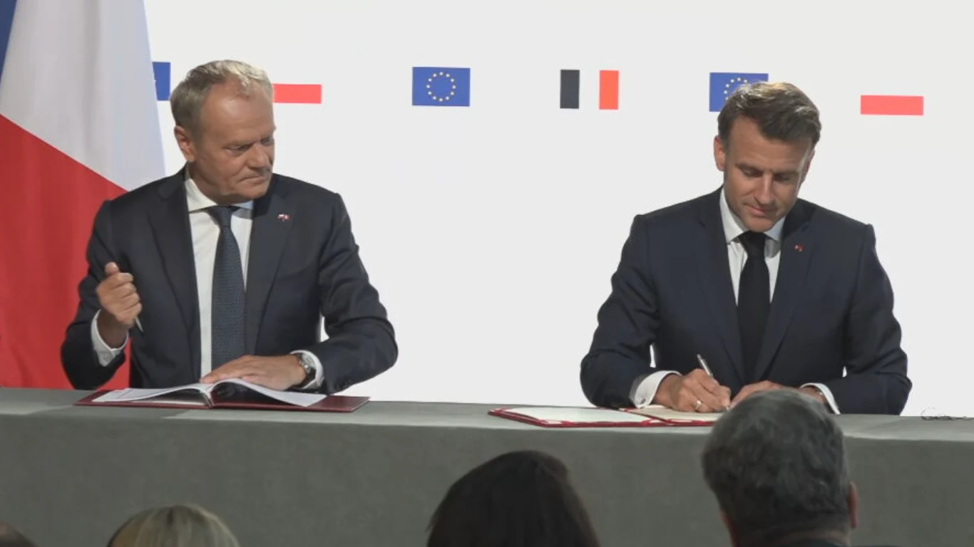 Polska i Francja podpisały traktat o współpracy i przyjaźni Dwaj mężczyźni w garniturach siedzą przy stole, podpisując dokumenty. Obaj są skupieni na podpisywaniu, a za nimi widoczne są flagi Unii Europejskiej i innych państw na białym tle.