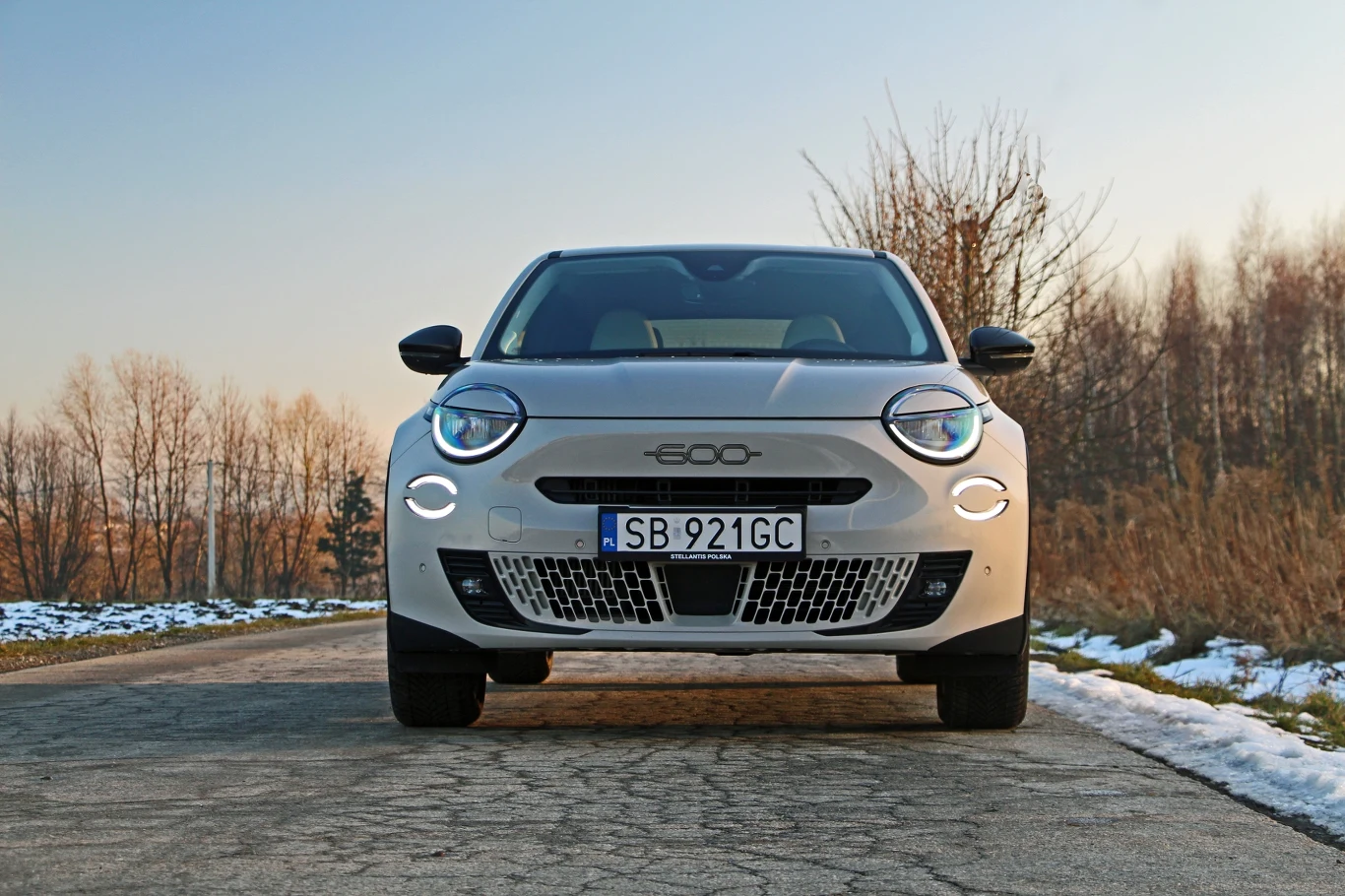 Samochód osobowy marki Fiat 500 ustawiony na wiejskiej drodze w zimowym krajobrazie. Widoczne są reflektory, tablica rejestracyjna oraz pokrywa silnika. W tle gołe drzewa i resztki śniegu.