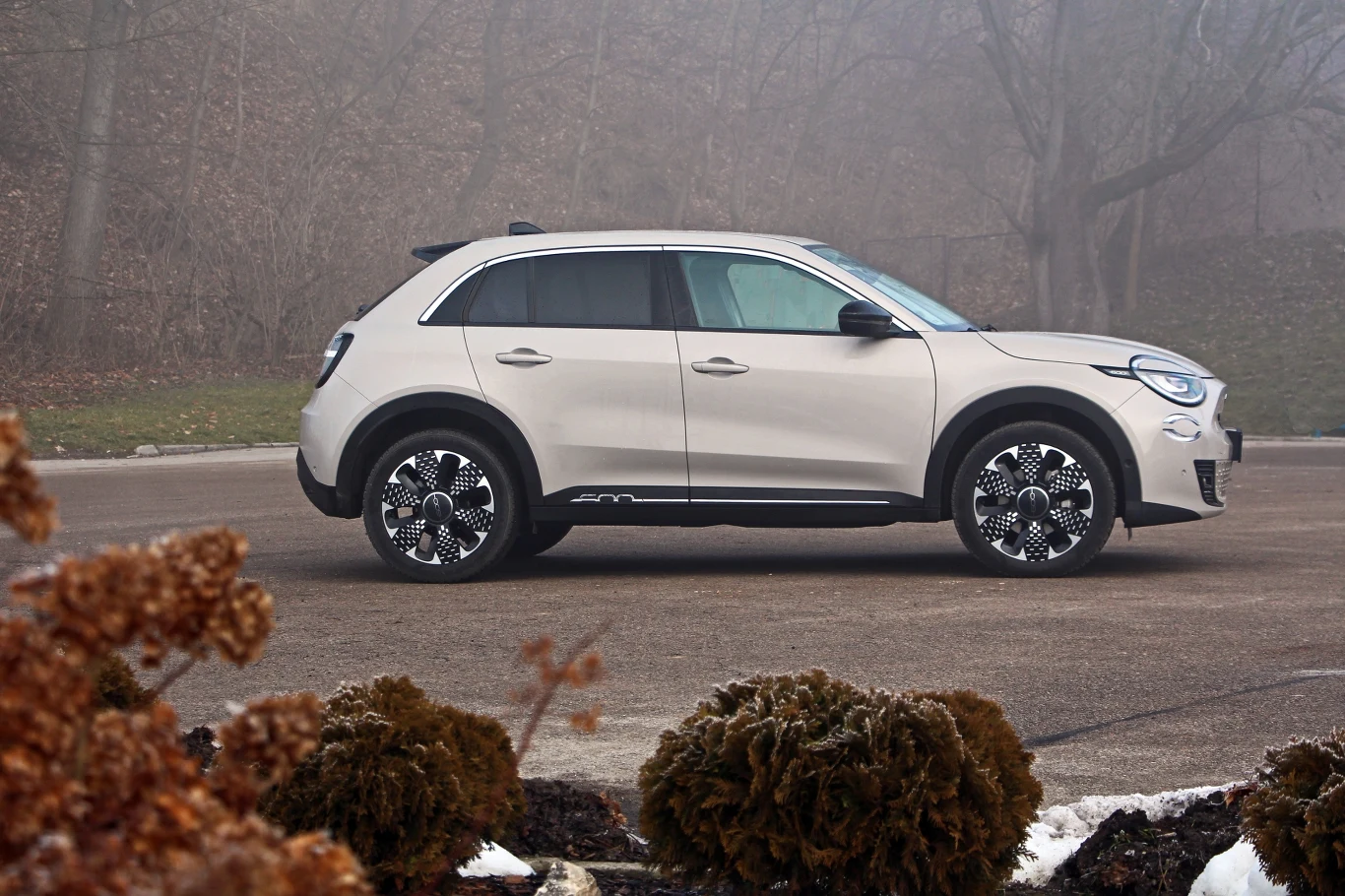 Nowoczesny samochód typu SUV w jasno-szarym kolorze stoi zaparkowany na asfaltowej drodze w otoczeniu zamglonego, jesiennego krajobrazu. W tle widać drzewa bez liści, a na pierwszym planie fragment niskiej roślinności.