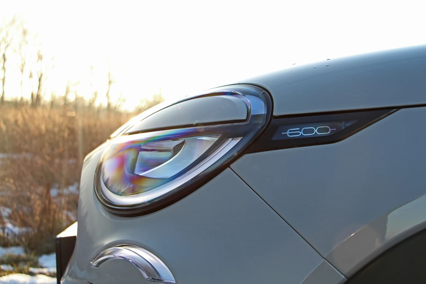 Zbliżenie na przedni reflektor samochodu, skupiające uwagę na precyzyjnych liniach i wyrazistym designie. W oddali widać elementy przyrody, co dodaje zdjęciu naturalnego tła i kontrastu.