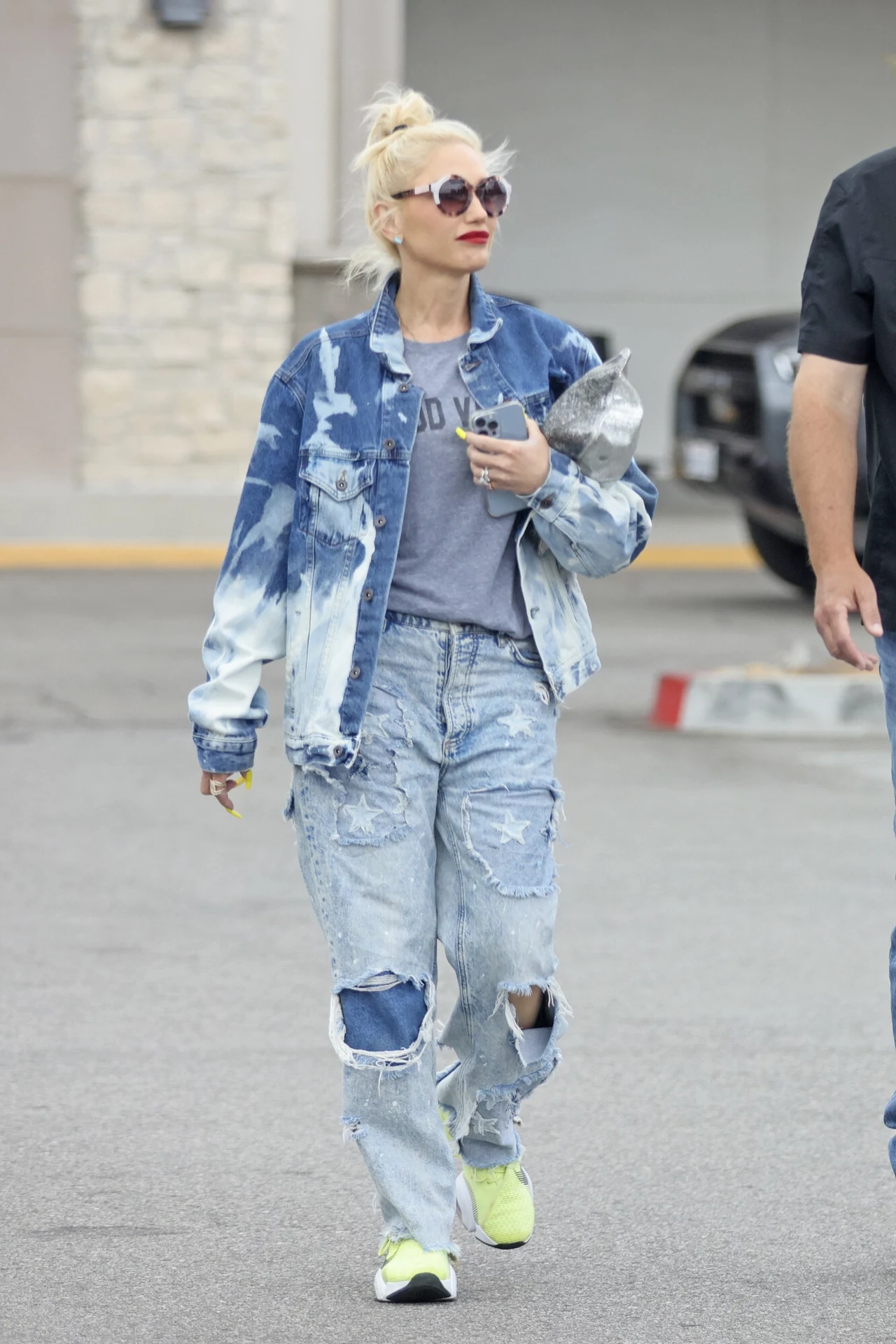 Gwen stefani w dżinsowym total looku Kobieta w rozjaśnionych jeansach z dziurami oraz jeansowej kurtce, trzymająca w ręce telefon, idzie po ulicy. Na nogach ma jasne sportowe buty, a na twarzy stylowe okulary przeciwsłoneczne. W tle fragment samochodu i budynku.