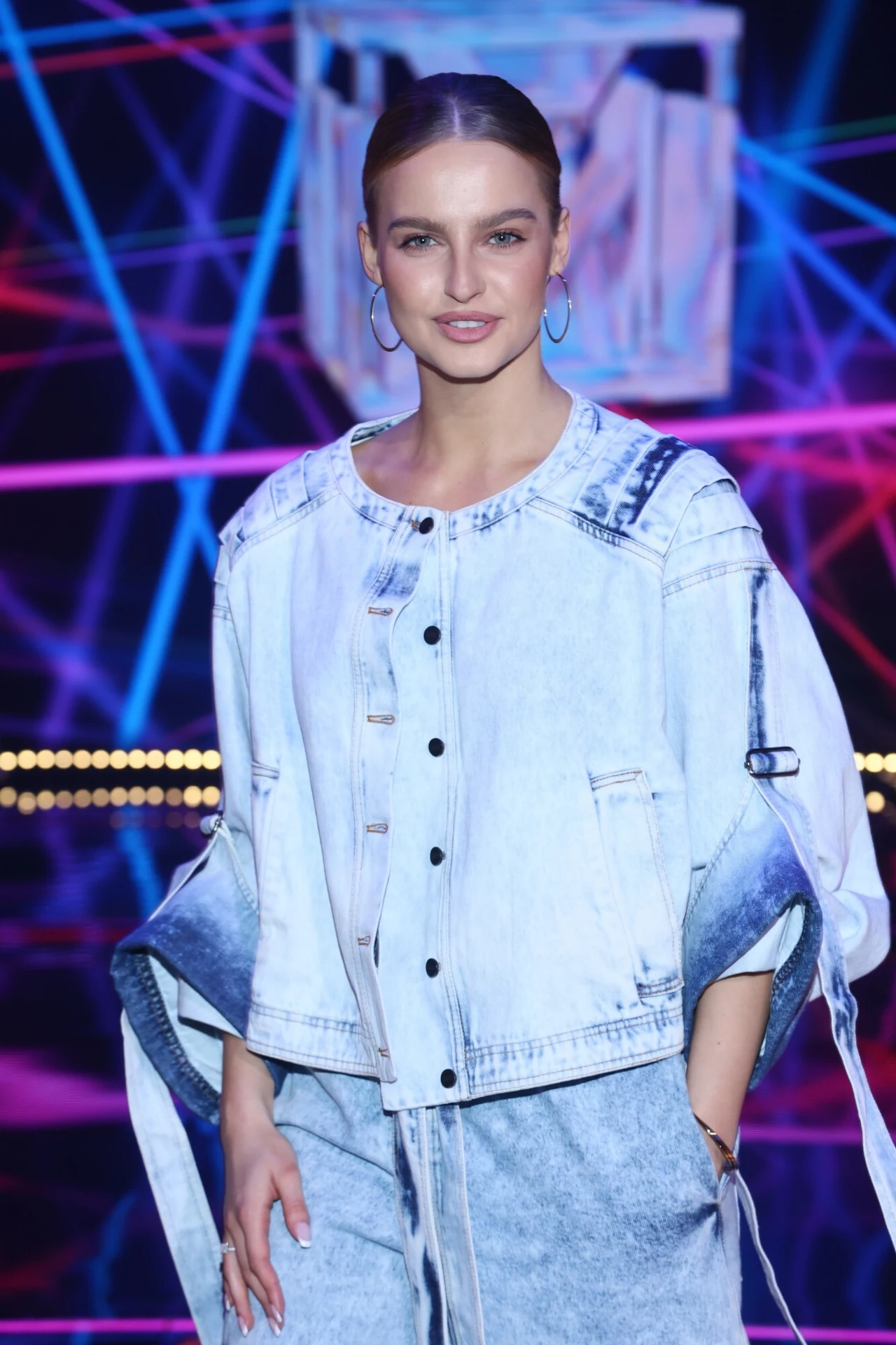 Karolina Pisarek podczas finału programu "Top model" Osoba ubrana w niebieski strój jeansowy stoi na tle kolorowych, świetlnych wzorów, uśmiechając się delikatnie.