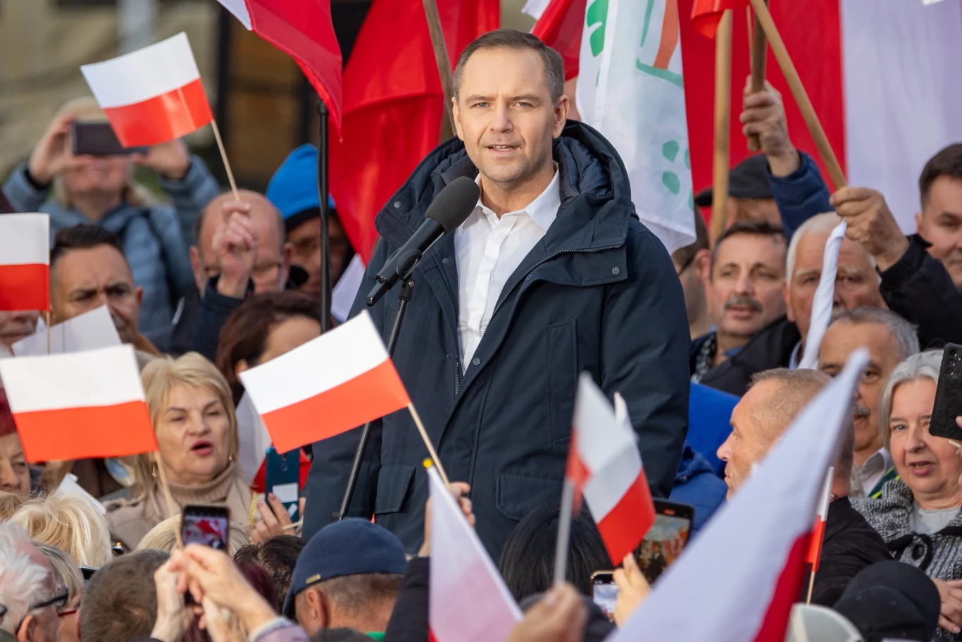 Karol Nawrocki od tygodnia musi tłumaczyć się z okoliczności nabycia mieszkania w Gdańsku Mężczyzna przemawia do tłumu ludzi na wiecu, trzymając mikrofon. W tle widać liczne polskie flagi, co sugeruje, że wydarzenie ma charakter patriotyczny lub polityczny.