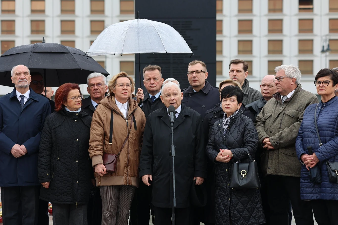 Kaczyński wydał oświadczenie podczas miesięcznicy smoleńskiej Grupa osób stojących razem przed mikrofonem, prawdopodobnie uczestników wydarzenia politycznego. W tle widoczna jest architektura budynków. Osoby są ubrani w płaszcze, co sugeruje chłodniejszą porę roku.