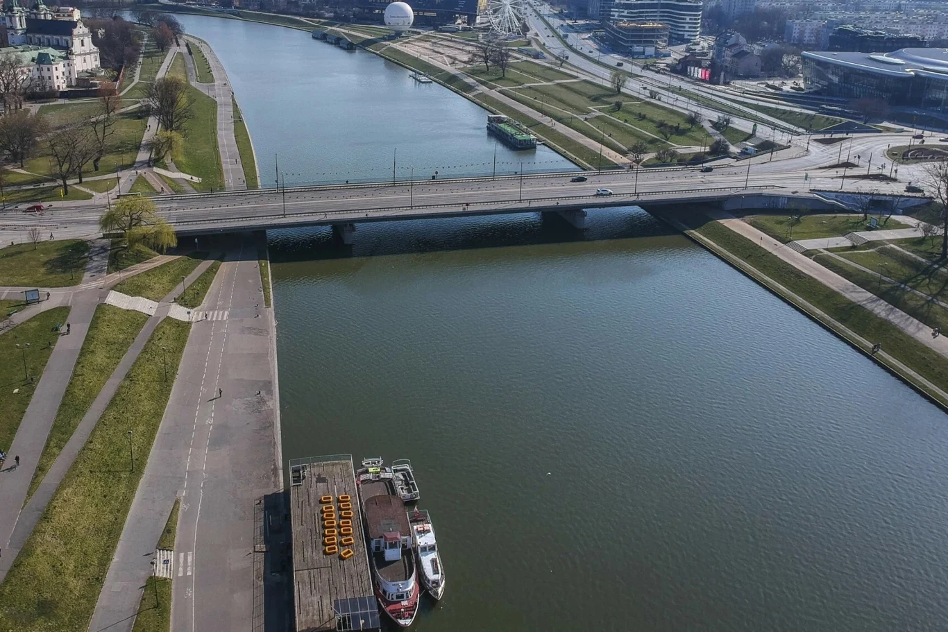 Remont mostu Grunwaldzkiego w Krakowie potrwa kilka miesięcy Most nad spokojną rzeką w mieście, na brzegach widoczne są zielone tereny oraz zabudowania miejskie w tle. Na rzece cumuje łódź przy nabrzeżu.