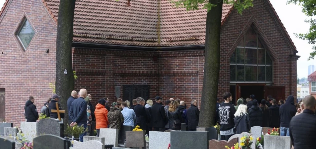 Pogrzeb Joki Grupa ludzi stojąca przed ceglaną kaplicą na cmentarzu, gdzie odbywa się ceremonia pogrzebowa. Kilka osób trzyma kwiaty. Z tyłu widoczne są nagrobki oraz roślinność, co podkreśla atmosferę skupienia i żalu.