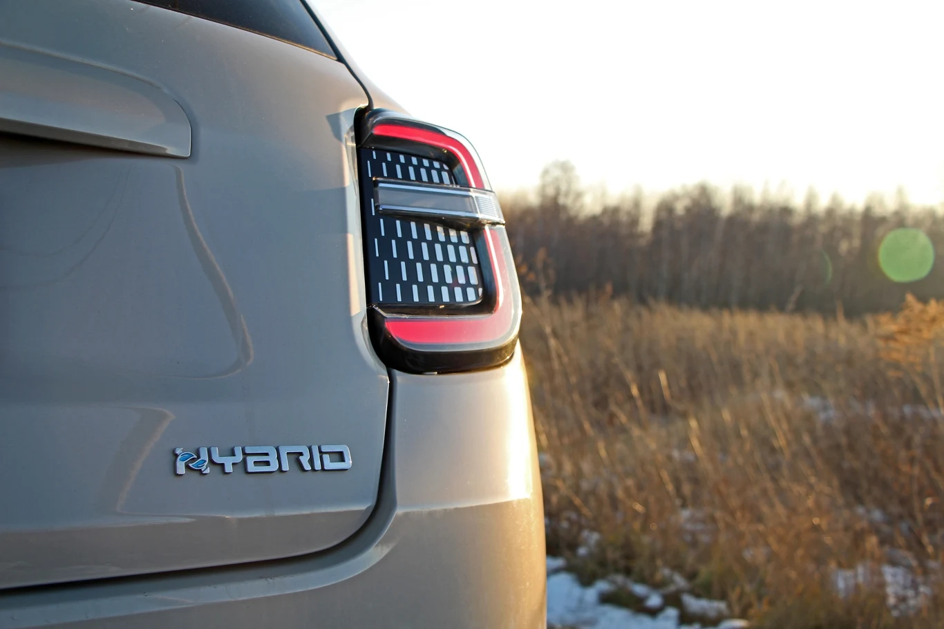 Tylny fragment samochodu hybrydowego na otwartej przestrzeni, z widocznym logo 'HYBRID' oraz nowoczesnymi światłami tylnymi, w tle trawiasta łąka i drzewa.
