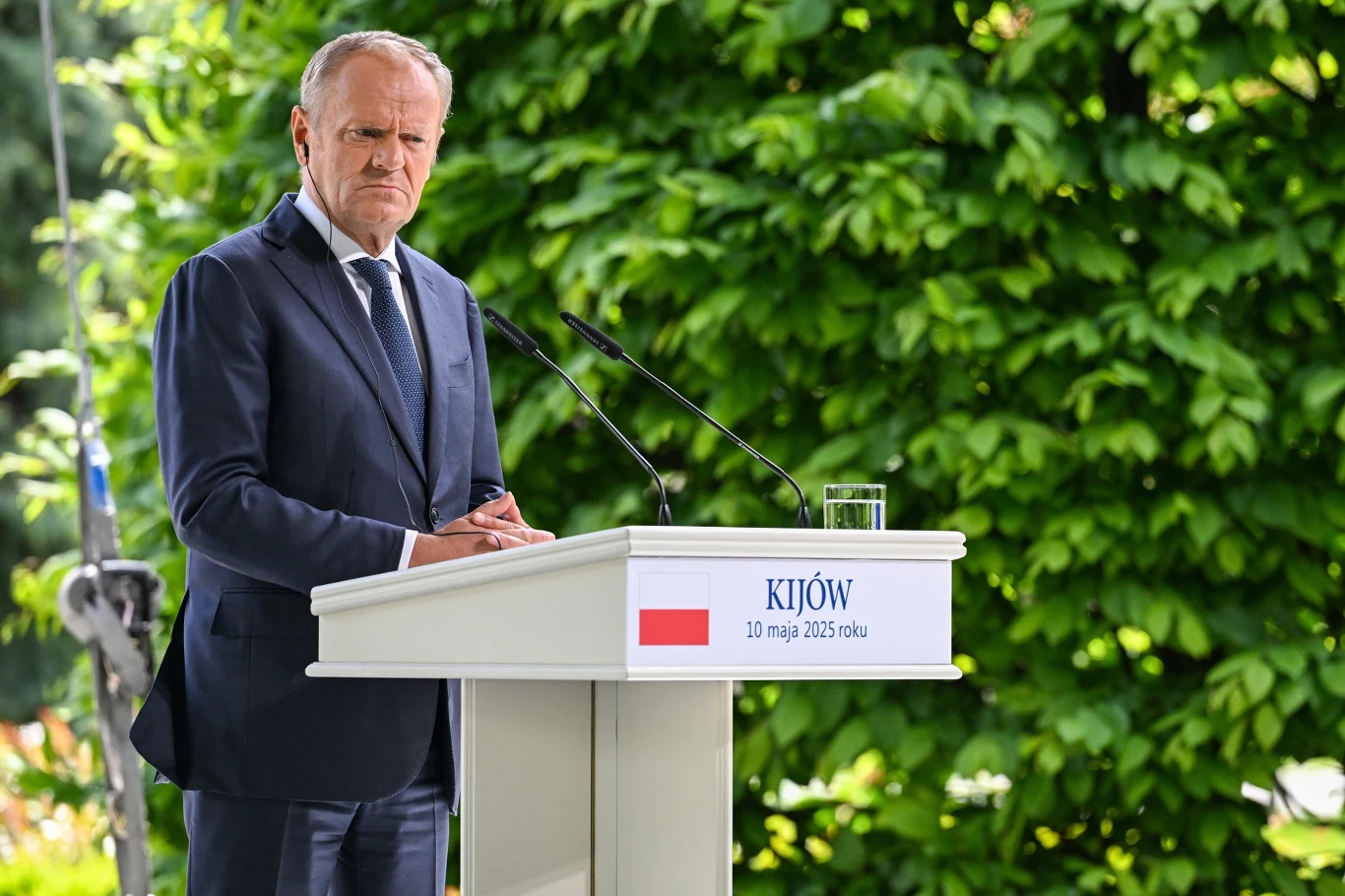 Premier Donald Tusk podczas konferencji w Kijowie Mężczyzna w garniturze przemawiający przy mównicy z napisem 'Kijów, 10 maja 2023 roku' na tle zielonego ogrodu.