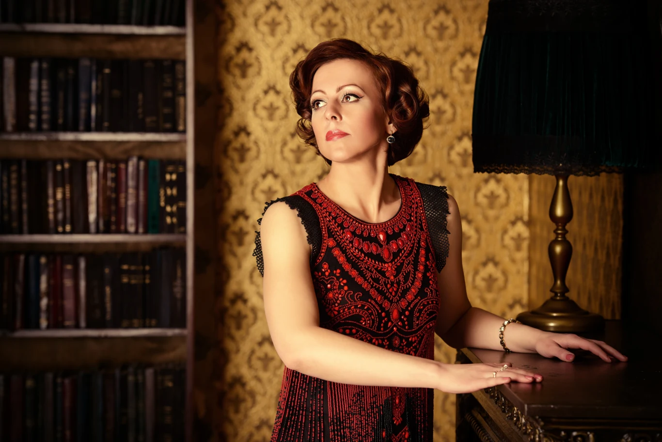 Co wydarzy się w najbliższych dniach? Kobieta o eleganckiej fryzurze retro, ubrana w bogato zdobioną suknię, pozuje w klasycznie urządzonym wnętrzu. Obok znajduje się ciemna lampa oraz półka z książkami, co dodaje scenie wyrafinowanego charakteru.