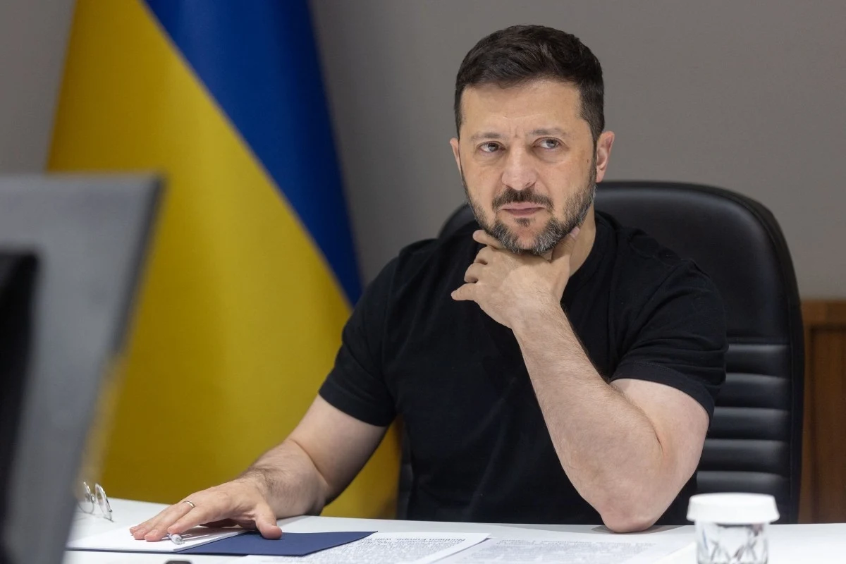 Wołodymyr Zełenski Mężczyzna w czarnym ubraniu siedzi przy stole z dokumentami, w tle widoczna flaga Ukrainy.