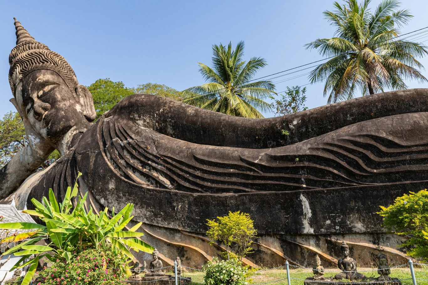 Leżący Budda to jeden z kluczowych motywów ikonograficznych w sztuce buddyjskiej (zdj. poglądowe) Leżący posąg Buddy otoczony bujną zielenią, z palmami w tle w słoneczny dzień.