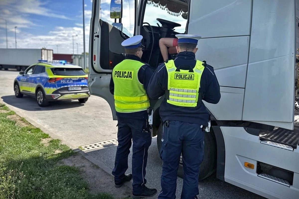 Policjanci kontrolowali czy kierowcy przestrzegają zakaz wyprzedzania przez ciężarówki na drogach szybkiego ruchu Policjanci w odblaskowych kamizelkach przeprowadzają kontrolę kierowcy ciężarówki. W tle stoi radiowóz policyjny, a scena ma miejsce w terenie otwartym.