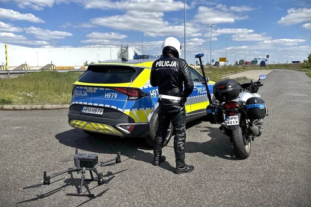 Policja coraz częściej korzysta z dronów do kontrolowania uczestników ruchu drogowego Policjant w kombinezonie motocyklowym stoi obok policyjnego samochodu i motocykla na otwartej drodze. Na ziemi leży dron gotowy do startu. Niebo jest jasne i bezchmurne, tworząc spokojne tło dla sceny.