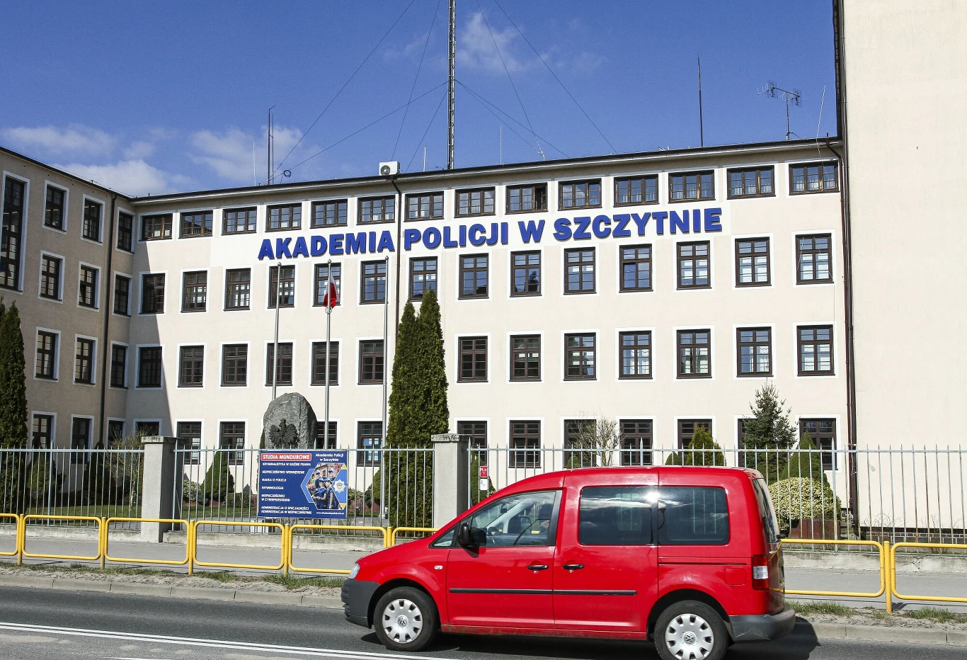 Masowe zatrucie w Akademii Policji w Szczytnie Budynek Akademii Policji w Szczytnie z dużym napisem na fasadzie. Przed budynkiem stoi czerwona furgonetka, a całość jest otoczona metalowym ogrodzeniem. Widoczna jest również flaga Polski.