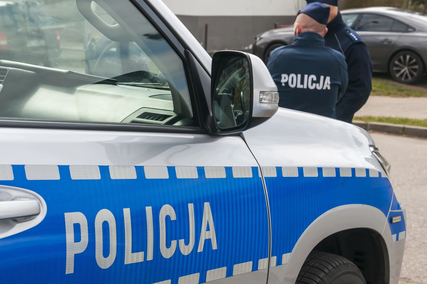 Małopolskie. 48-latek usłyszał dwa zarzuty. Jest podejrzany o pozbawienie życia matki, zdj. ilustracyjne Radiowóz policyjny na pierwszym planie z dwoma policjantami w tle. Samochód jest oznakowany, a funkcjonariusze ubrani są w mundury. Scena odbywa się prawdopodobnie w mieście, na poboczu drogi.
