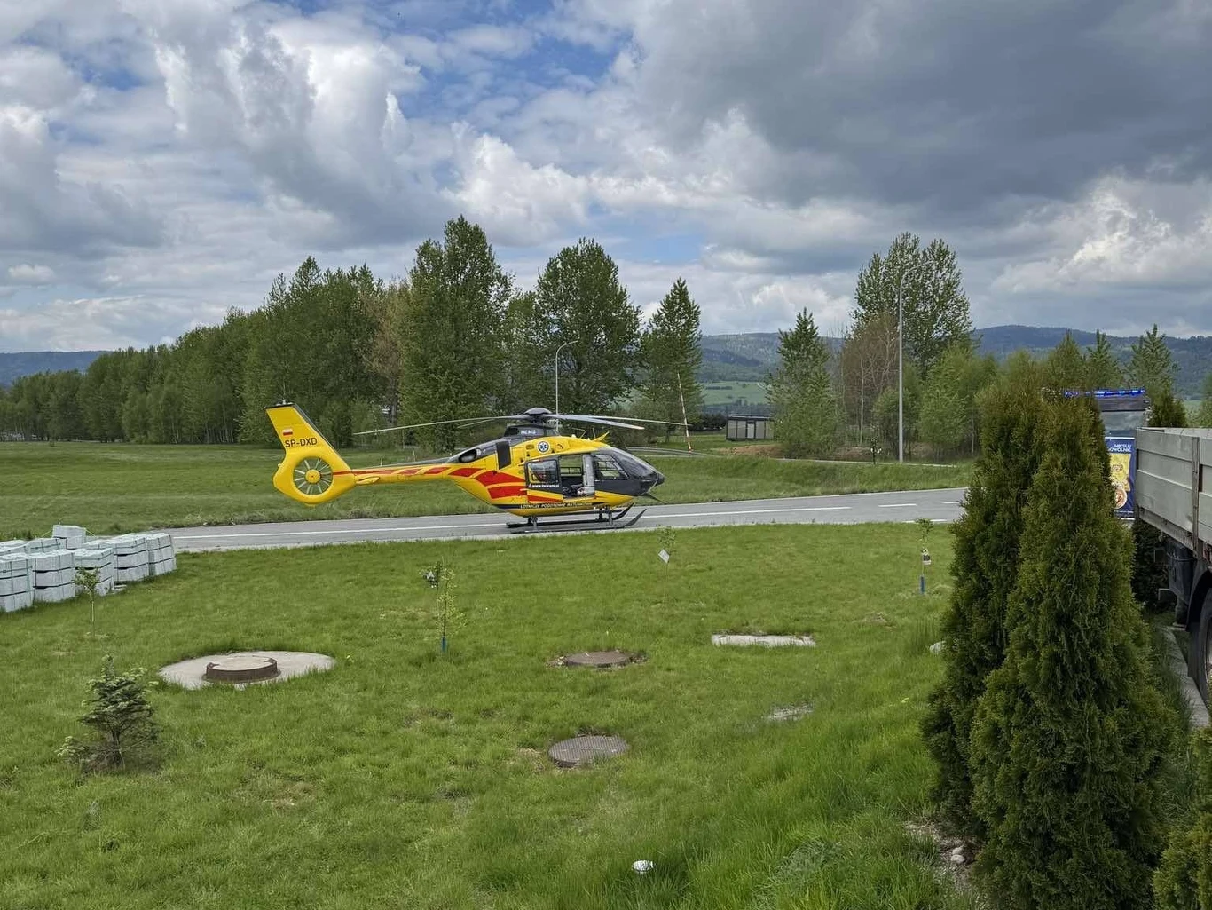 Wypadek w Nowym Targu. Nie żyje spadochroniarz Żółty helikopter ratunkowy lądujący na trawniku w otoczeniu drzew i gór w tle. Na pierwszym planie widoczne są drzewa i fragment pojazdu ciężarowego po prawej stronie.