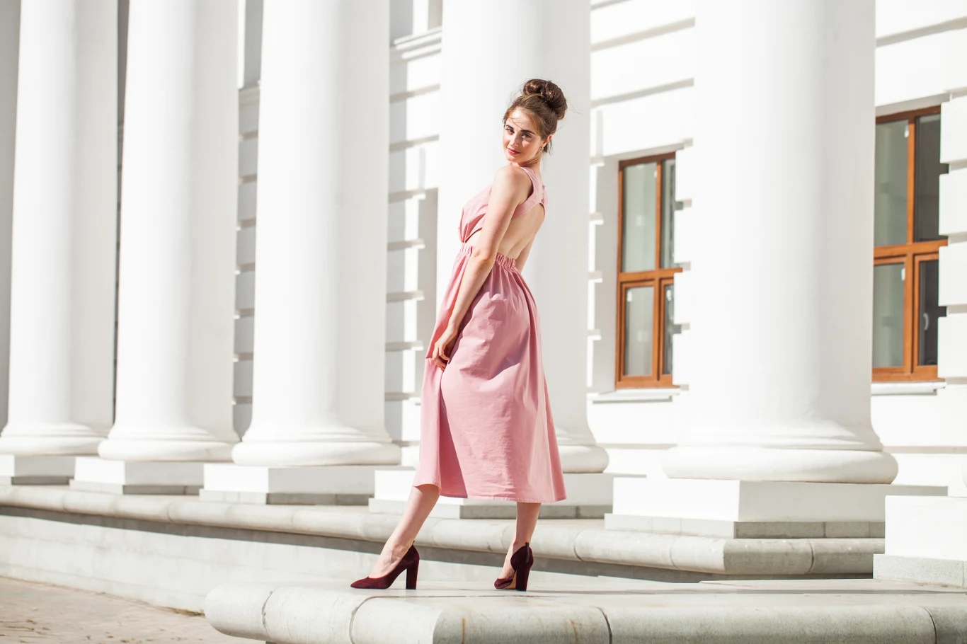 Wybierają buty na wesele warto pamiętać, że mają być eleganckie, ale też wygodnie. Kobieta w różowej sukience stoi na tle białych kolumn budynku. Jej włosy są upięte, a na nogach ma czerwone buty na wysokim obcasie. Światło podkreśla elegancję i szyk stylizacji.