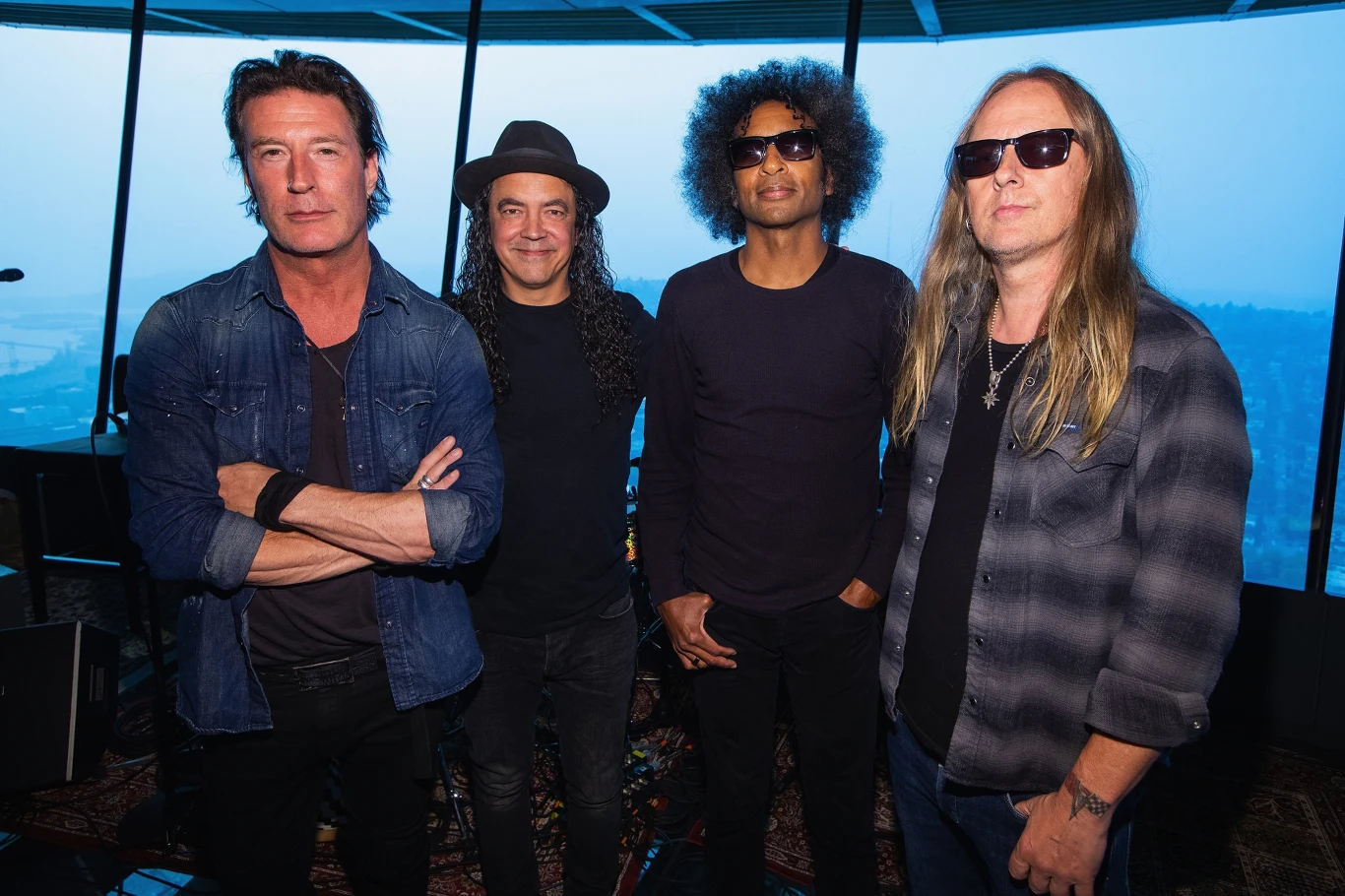 Sean Kinney, Mike Inez, William DuVall i Jerry Cantrell (Alice In Chains) w 2018 r. Czterech mężczyzn stoi obok siebie, wszyscy patrzą w obiektyw aparatu. Każdy z nich ma swój unikalny styl ubioru. W tle widać duże okna, przez które wpada naturalne światło.