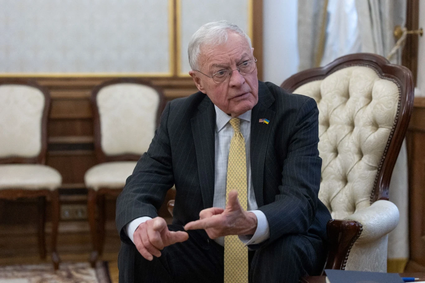 Specjalny wysłannik USA ds. Ukrainy gen. Keith Kellogg Mężczyzna w garniturze z krawatem siedzący na eleganckim krześle w sali z drewnianymi panelami na ścianach, obok stoją inne krzesła. W tle widać zasłony i fragment okna.