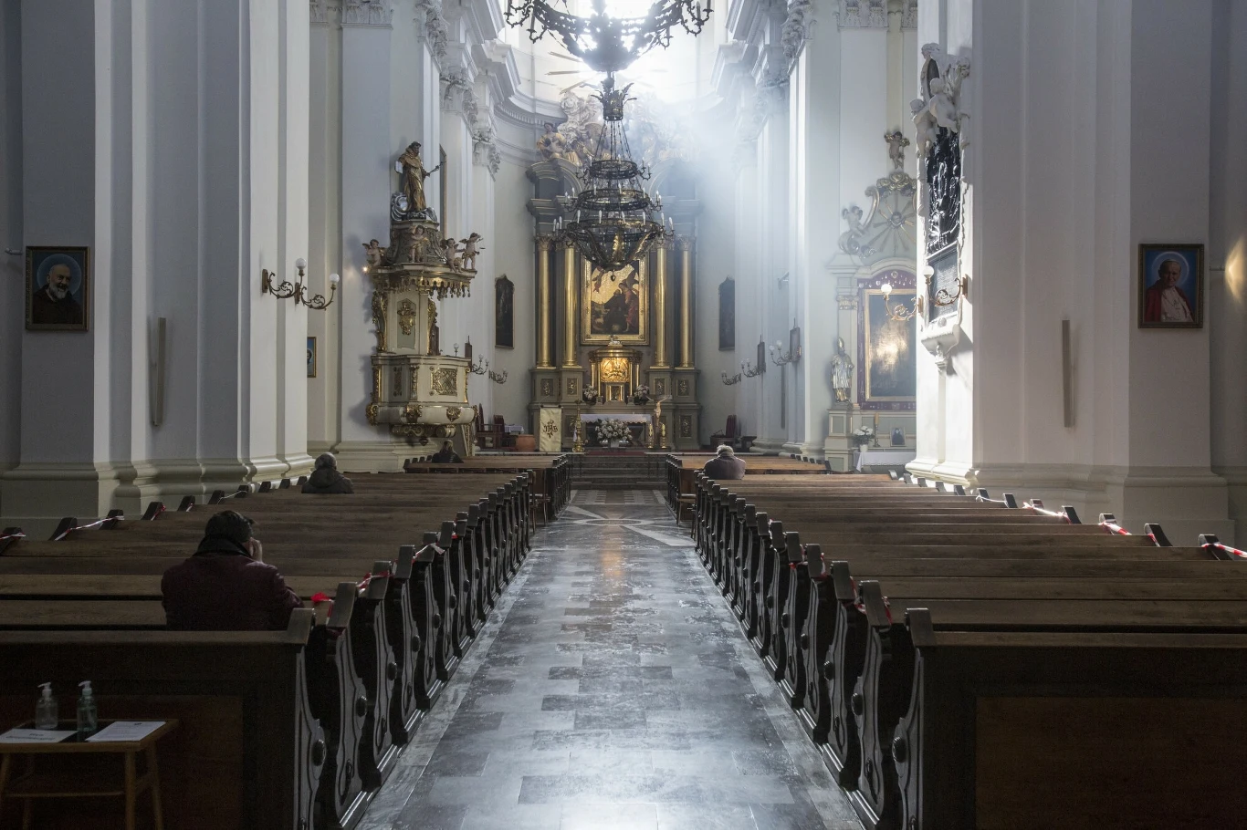 Zdjęcie ilustracyjne Wnętrze dużego, historycznego kościoła z wysokimi, jasnymi ścianami i drewnianymi ławkami. Światło wpadające przez okno oświetla centralną nawę, w tle widać ołtarz. Kilka osób siedzi w milczeniu w ławkach.