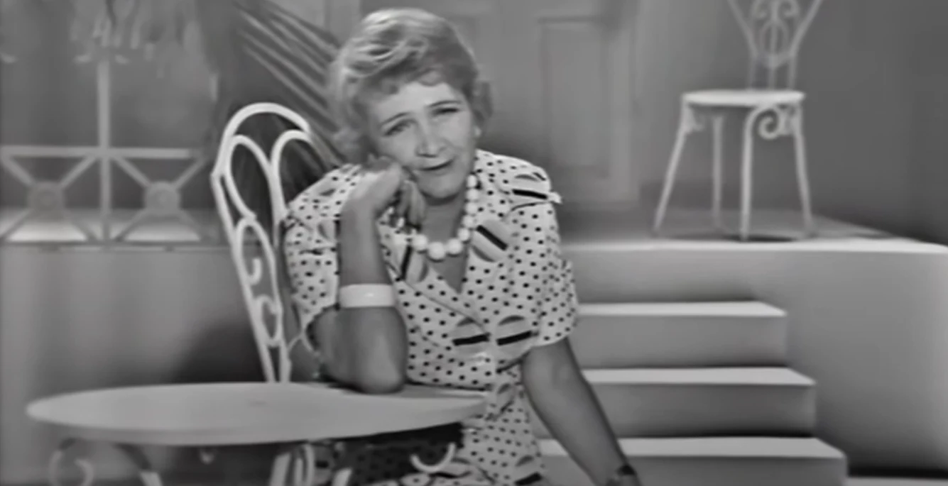 Marta Mirska Starsza kobieta w sukience w groszki siedzi przy stoliku kawiarnianym. Sceneria jest retro, z wyraźnie stylizowanymi krzesłami i schodami w tle, co dodaje klimatu dawnych czasów.