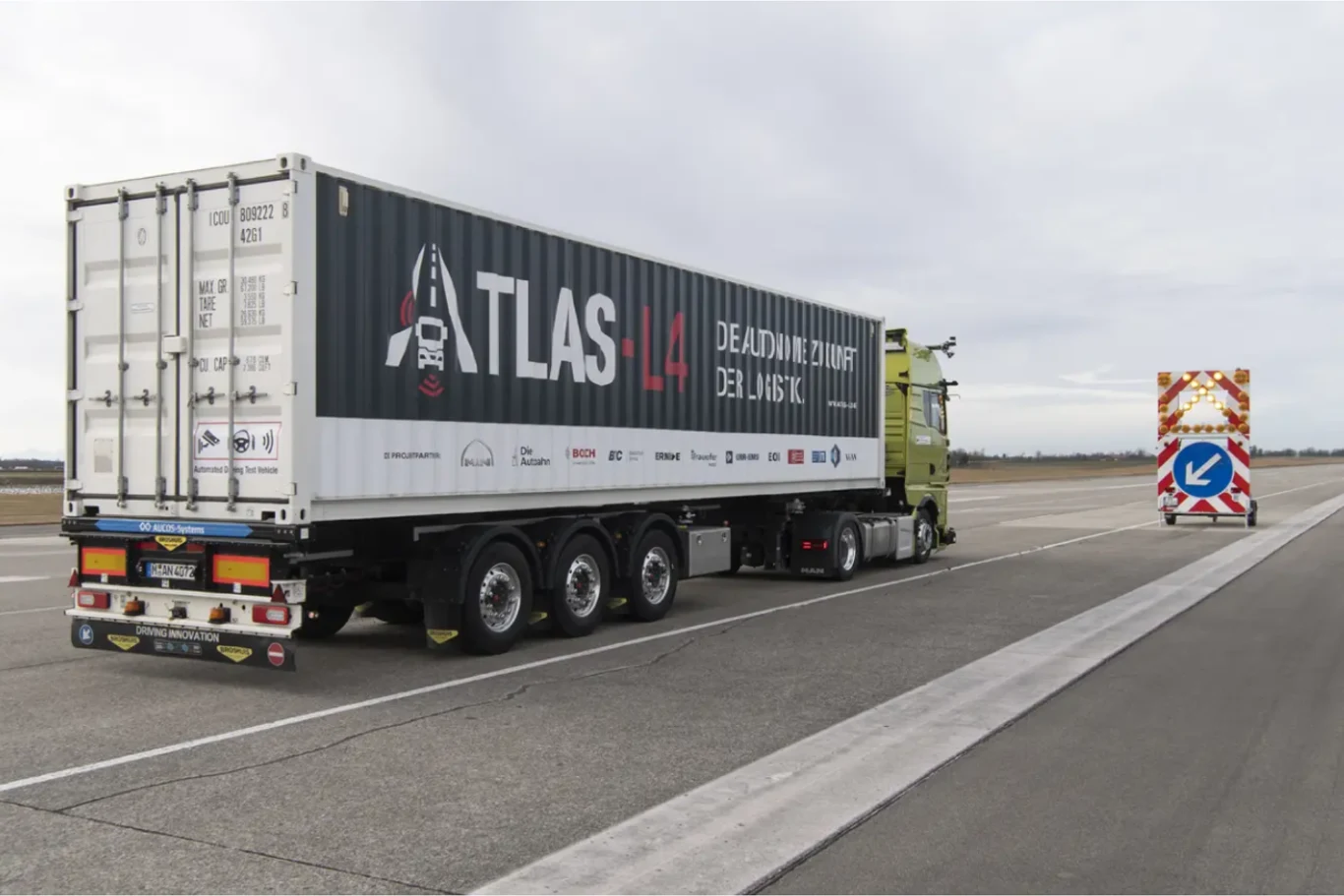 Ciężarówka z kontenerem oznaczonym ATLAS L4 na pasie drogi. Kontener jest częścią infrastruktury logistycznej, a wokół znajdują się znaki drogowe kierujące ruchem.