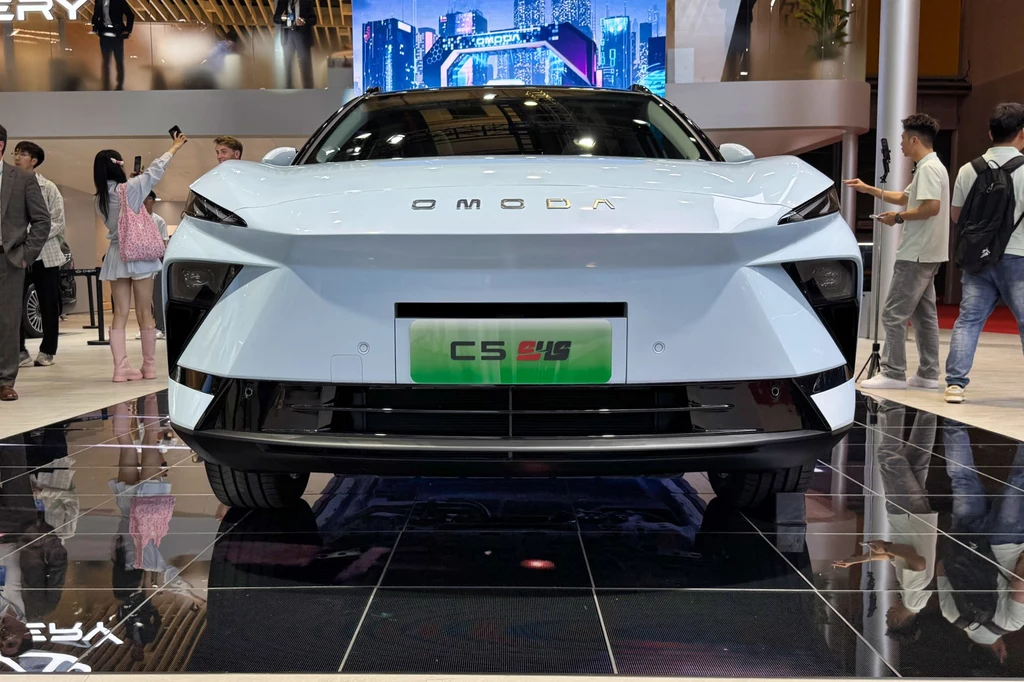 Obecnie w Omodzie 5 dominuje ogromny gril. W nowym modelu zredukowano go do wąskiego paska w dolnej części zderzaka Nowoczesny samochód elektryczny Omoda C5 EV w kolorze białym prezentowany na wystawie motoryzacyjnej, otoczony przez odwiedzających.