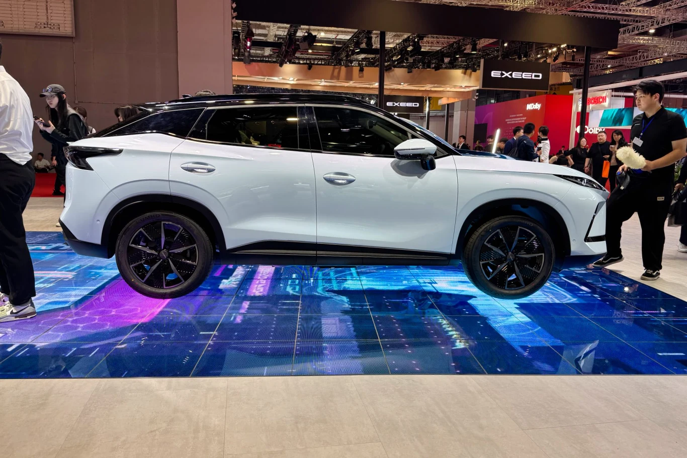 Biały samochód SUV na podświetlonej platformie z efektami świetlnymi, otoczony przez widzów na wystawie samochodowej.