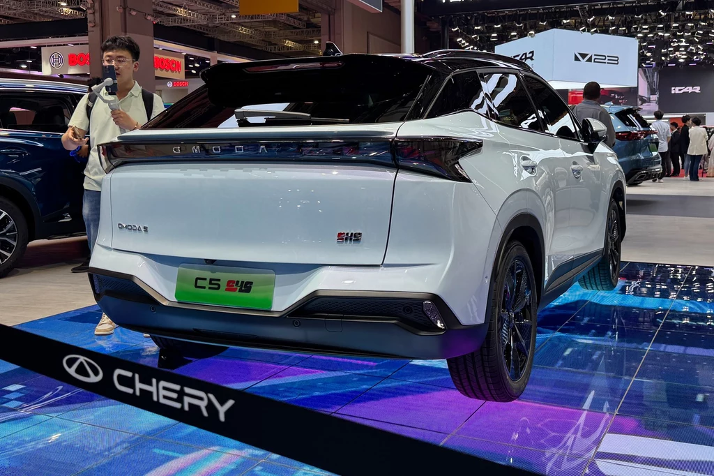 W tylnej części nadwozia pojawił się pas świetlny łączący obie lampy Samochód Chery w kolorze białym na tle wystawy motoryzacyjnej, widoczny tył z wyraźnymi oznaczeniami marki. Podświetlona podłoga dodaje nowoczesnego wyglądu.