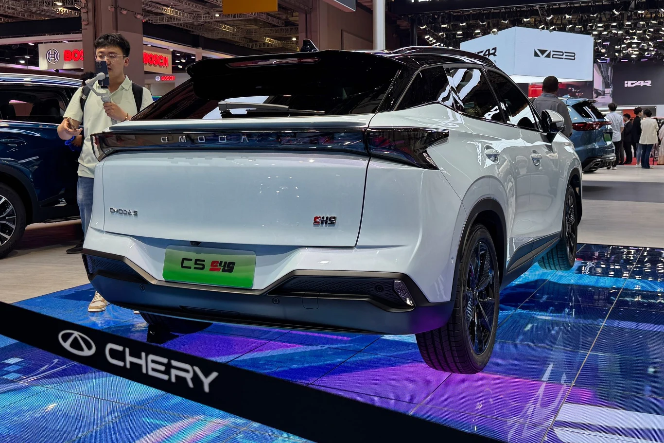 Samochód Chery w kolorze białym na tle wystawy motoryzacyjnej, widoczny tył z wyraźnymi oznaczeniami marki. Podświetlona podłoga dodaje nowoczesnego wyglądu.