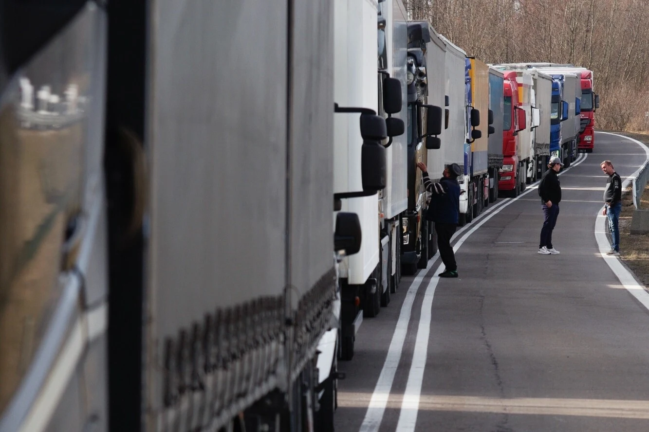 Przedsiębiorcy z branży transportowej zablokują granicę. Sąd unieważnił decyzję wójta. Ciężarówki ustawione w długiej kolejce na drodze, widać kilku kierowców stojących obok pojazdów, rozciągająca się na zakręcie asfaltowa droga otoczona przez drzewa.