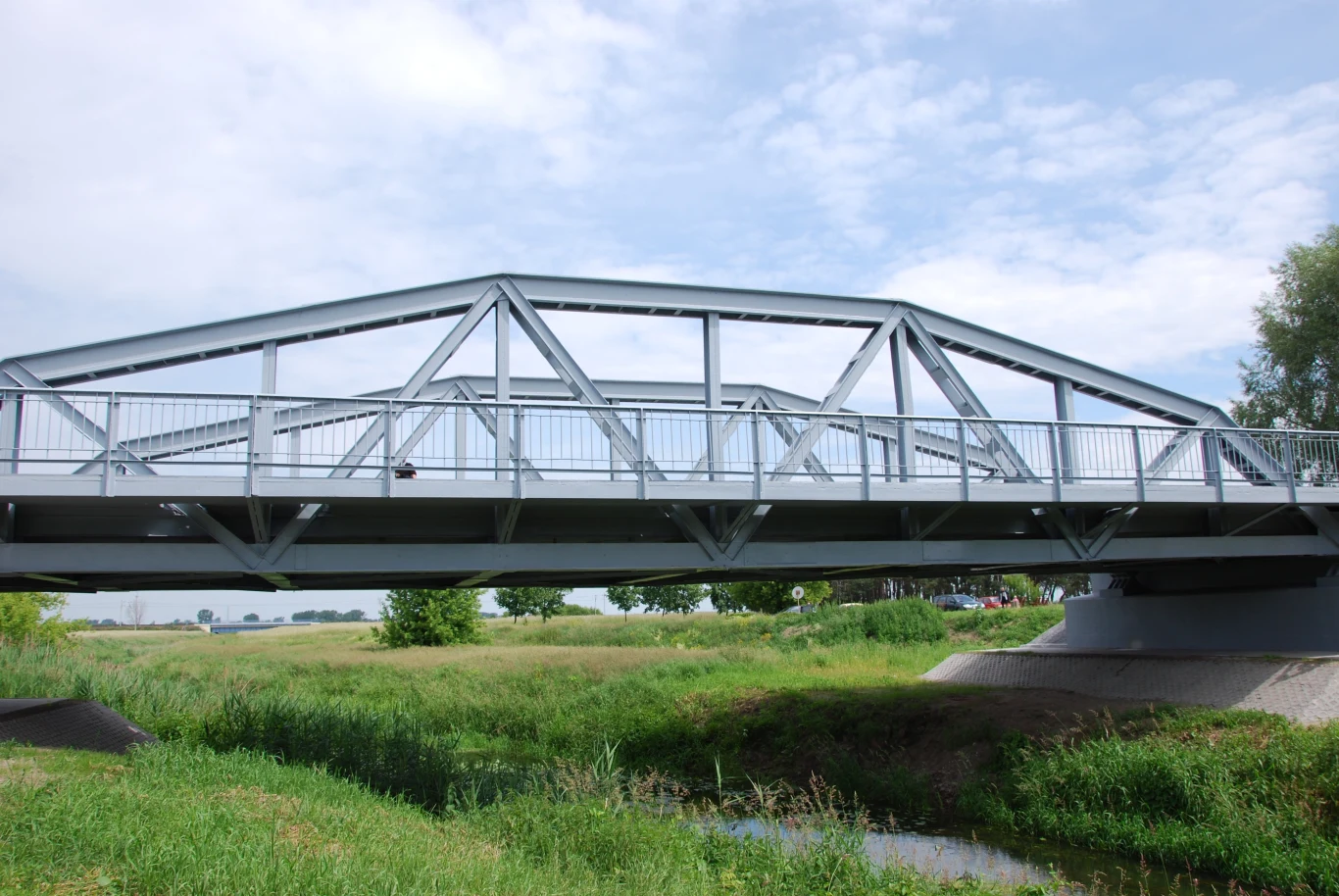 Most w Maurzycach to wyjątkowa konstrukcja, która pozbawiona jest charakterystycznych nitów. Stalowy most nad zieloną doliną, otoczony trawami i drzewami pod błękitnym niebem z delikatnymi chmurami.