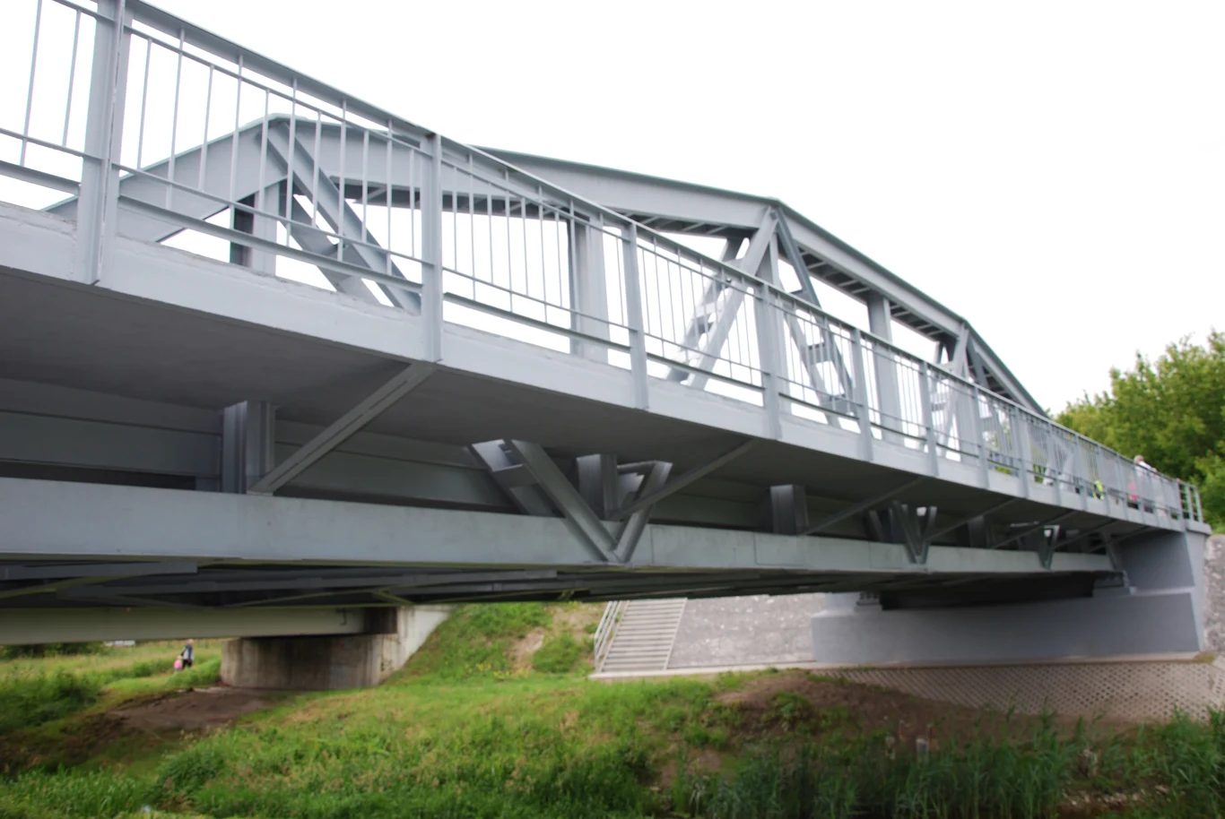 Masa mostu dzięki spawaniu zmniejszyła się z 70 do 56 ton. Most stalowy o strukturze kratownicowej, rozciągający się nad rzeką w spokojnym otoczeniu z roślinnością. Widoczna jest betonowa konstrukcja wspierająca oraz zielone obszary wokół mostu.