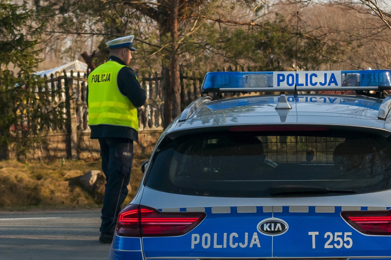 "Obywatelskie ujęcie" w Lipsku. Zabrał kobiecie kluczyki do samochodu Policjant w żółtej kamizelce odblaskowej stoi obok oznakowanego radiowozu marki Kia, z napisem 'Policja'. W tle widoczna jest zieleń i płot.