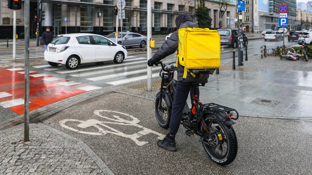 Kurier na rowerze elektrycznym z dużym, żółtym plecakiem na plecach porusza się po ścieżce rowerowej w miejskim otoczeniu. W tle widać samochody na ulicy i budynki. Mężczyzna ubrany w kurtkę i kaptur, co sugeruje chłodniejszą pogodę.