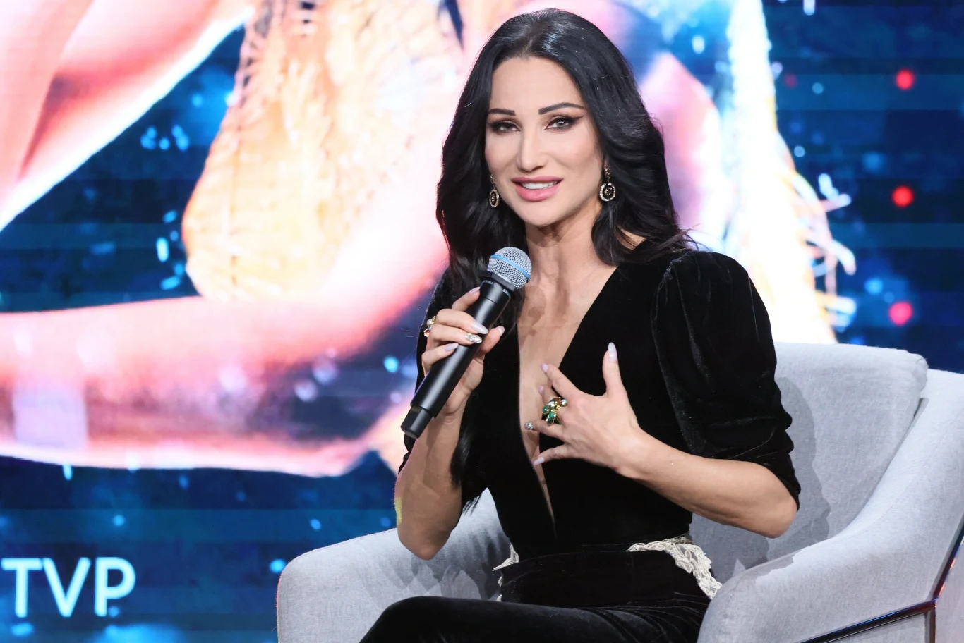 Justyna Steczkowska jest reprezentantką Polski na Eurowizji 2025 Kobieta o ciemnych włosach siedzi na krześle i trzyma w ręku mikrofon. Ma na sobie elegancką czarną suknię i biżuterię. Za nią znajduje się ekran z rozmytym tłem.