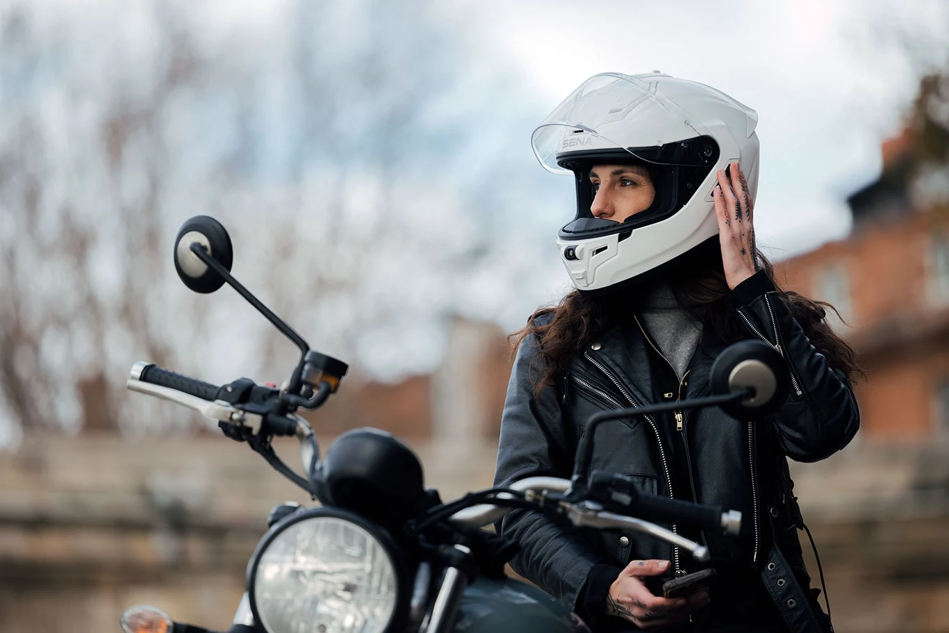 Osoba w białym kasku motocyklowym stoi przy motocyklu. Ubrana w czarną skórzaną kurtkę, patrzy w bok, trzymając rękę przy kasku.