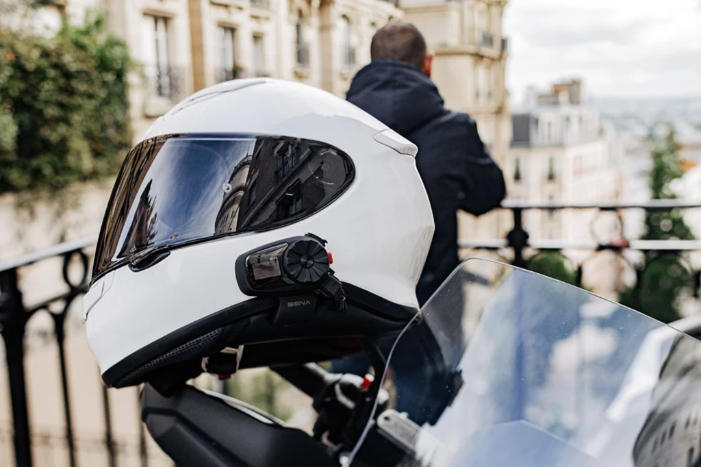Interkom marki Sena mocowany jest z boku kasku. Duże pokrętło służy do obsługiwania głównych funkcji. Biały kask motocyklowy z zamontowaną kamerą umieszczony na motocyklu, w tle nieostry widok miasta i sylwetka człowieka oparta o balkon.