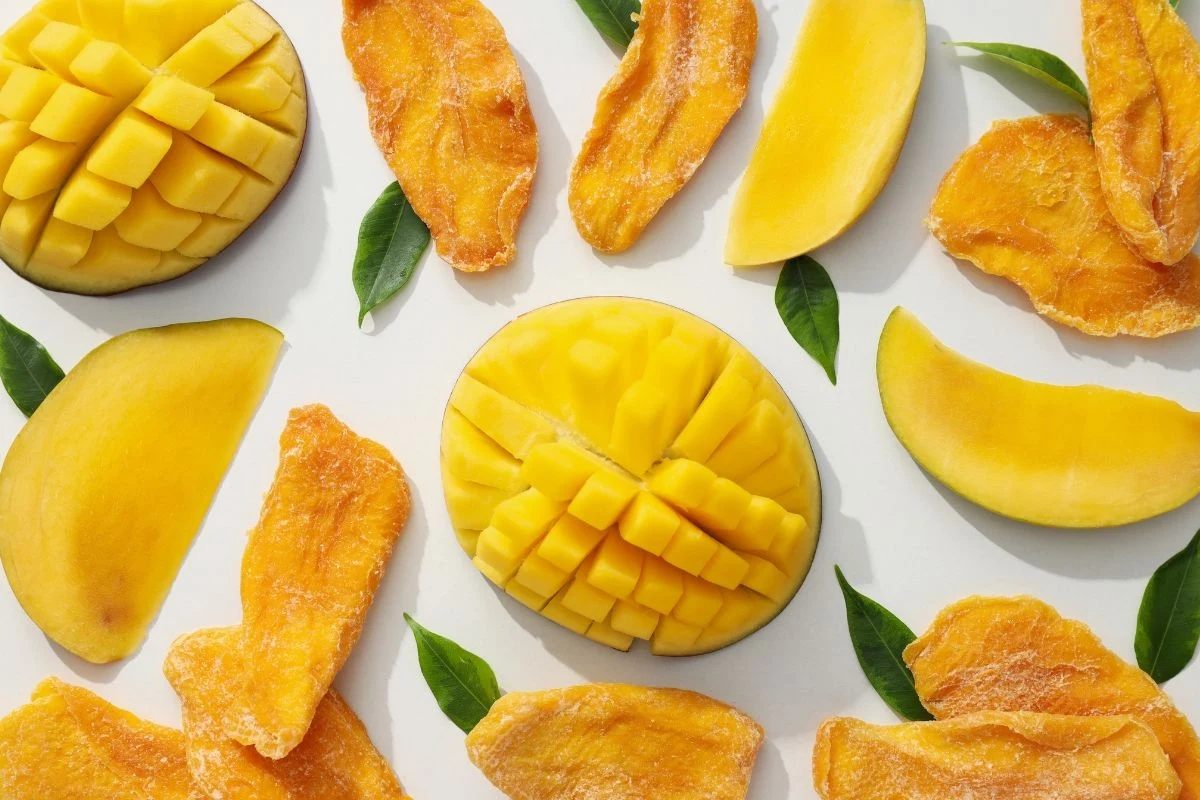 Plastry świeżego i suszonego mango ułożone symetrycznie na białym tle, tworząc atrakcyjny wzór. Soczyste kolory owoców kontrastują z zielonymi liśćmi, nadając kompozycji świeżości i egzotycznego wyglądu.