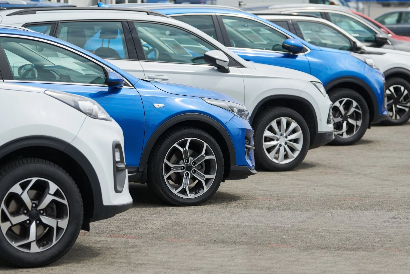 Samochody osobowe ustawione w rzędzie na parkingu, różne kolory i modele, wyraźnie widoczne detale nadwozia i kół.