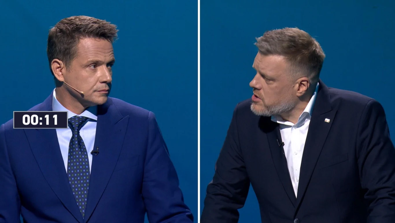 Rafał Trzaskowski i Adrian Zandberg podczas debaty Dwóch mężczyzn w garniturach uczestniczy w debacie na niebieskim tle. Pierwszy mężczyzna nosi niebieski krawat, drugi ma białą koszulę bez krawata. Między nimi widoczny jest licznik czasu. Obaj wyglądają na zaangażowanych w dyskusję.