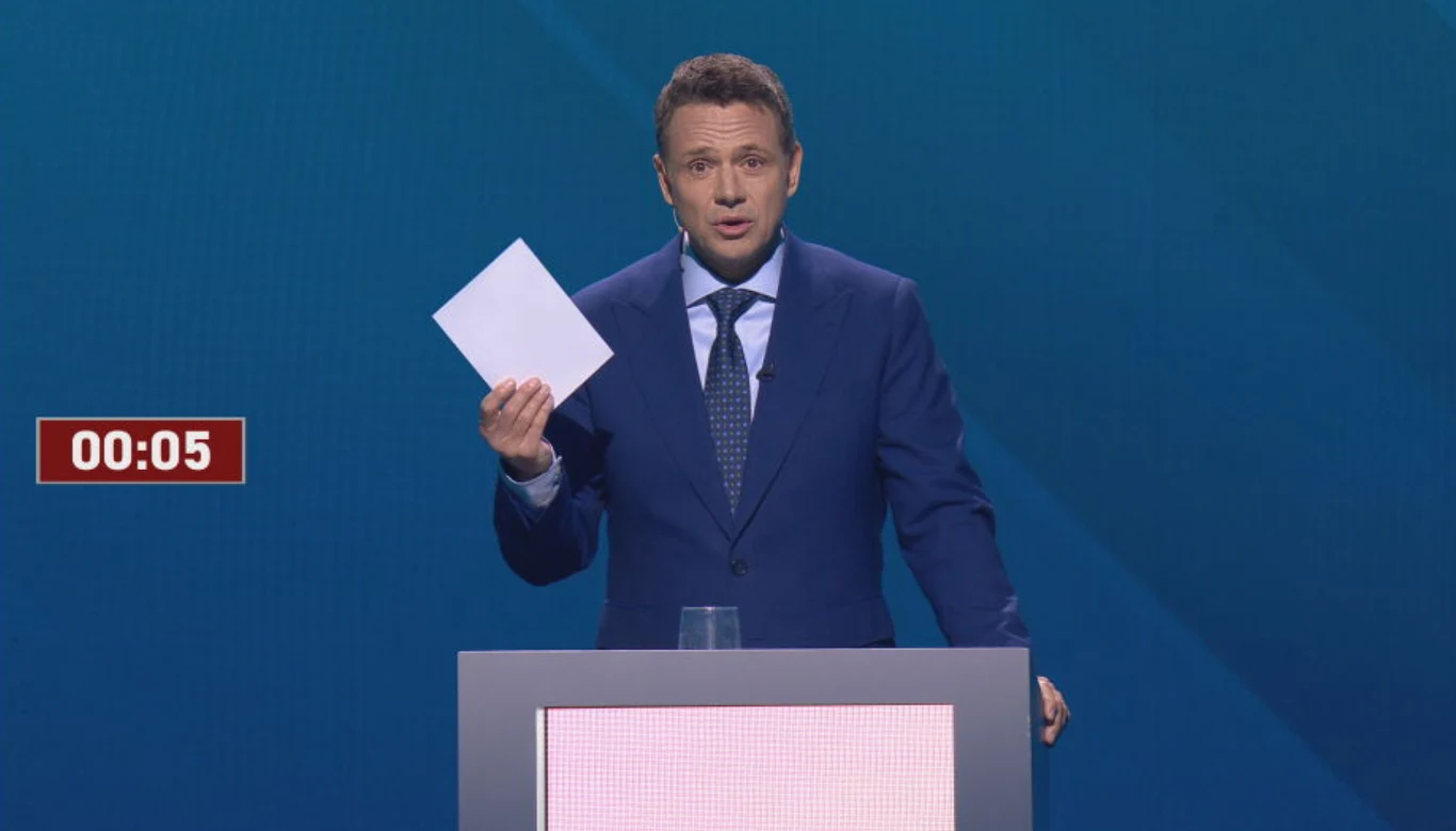 Wybory prezydenckie 2025. Na koniec debaty Rafał Trzaskowski wręczył Karolowi Nawrockiemu kopertę Mężczyzna w garniturze stoi przy mównicy, trzymając kartkę papieru z wyraźnym stoperem w tle wyświetlającym czas 00:05. Jego wyraz twarzy jest koncentrujący. Scena ma profesjonalny charakter i odbywa się na niebieskim tle.