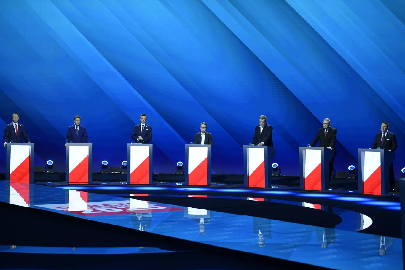 Lawina komentarzy po ostatniej debacie przedwyborczej Debata polityczna z udziałem siedmiu osób stojących za podium na tle niebieskiej sceny z oświetleniem. Każde podium ozdobione jest flagą w narodowych barwach. Cała scena jest nowoczesna i stylowa, utrzymana w tonacji niebieskiej.