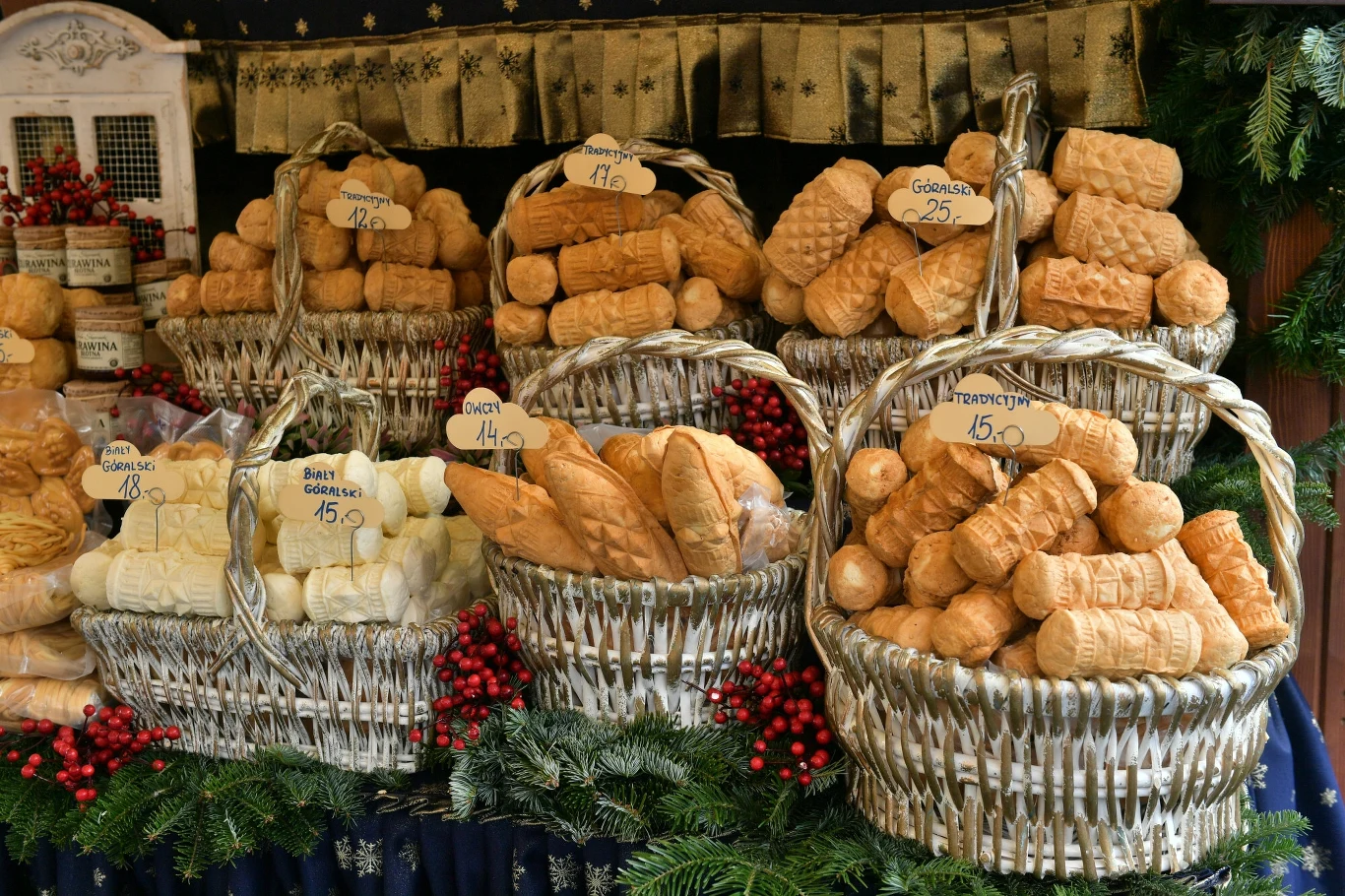 Stoisko z oscypkiem na jarmarku, pełne koszy z różnorodnymi serami w kształcie rolek, pośród dekoracji z gałązek iglastych i czerwonych jagód. Każdy kosz ma etykietę z ceną i oznaczeniem rodzaju sera.
