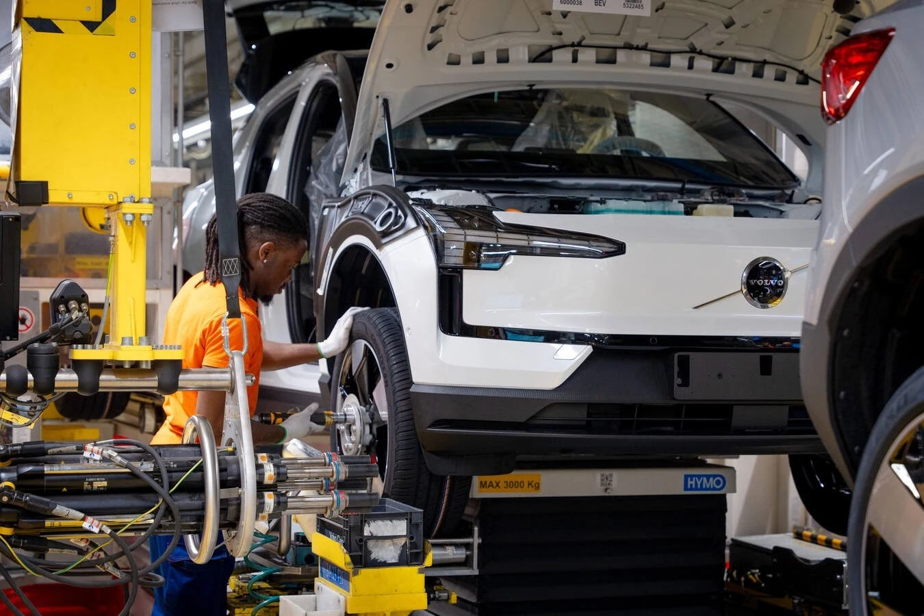 Volvo ogłosiło dużą akcję serwisową obejmująca łącznie 40 323 samochody. Pracownik montuje samochód w fabryce, pracujący nad kołem pojazdu. Otwarte maski ujawniają mechanizmy wewnętrzne, a wokół jest nowoczesne wyposażenie produkcyjne.