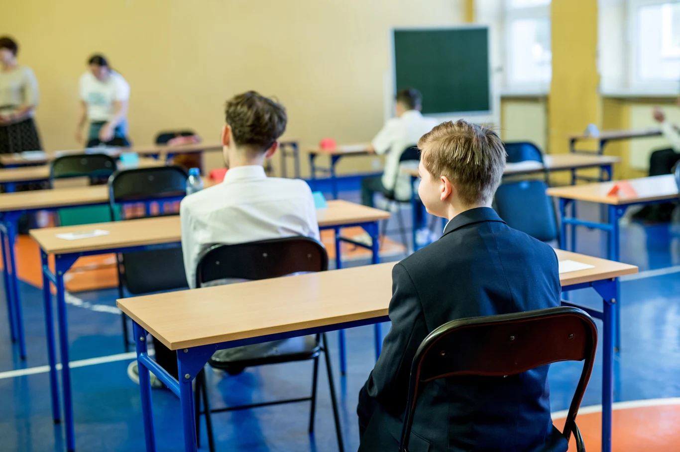 Egzamin ósmoklasisty wystartował Uczniowie siedzą przy stołach w sali egzaminacyjnej, ubrani formalnie. W tle tablica i kilka osób, które zajmują się organizacją sali.