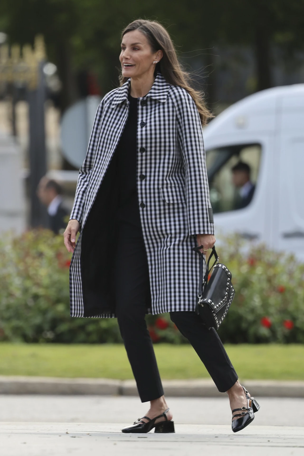 Królowa Letycja w eleganckiej stylizacji. W roli głównej: płaszcz w kratę Elegancko ubrana kobieta w biało-czarnym płaszczu, trzymająca czarną torebkę, spaceruje po ulicy. W tle widać białe auto.