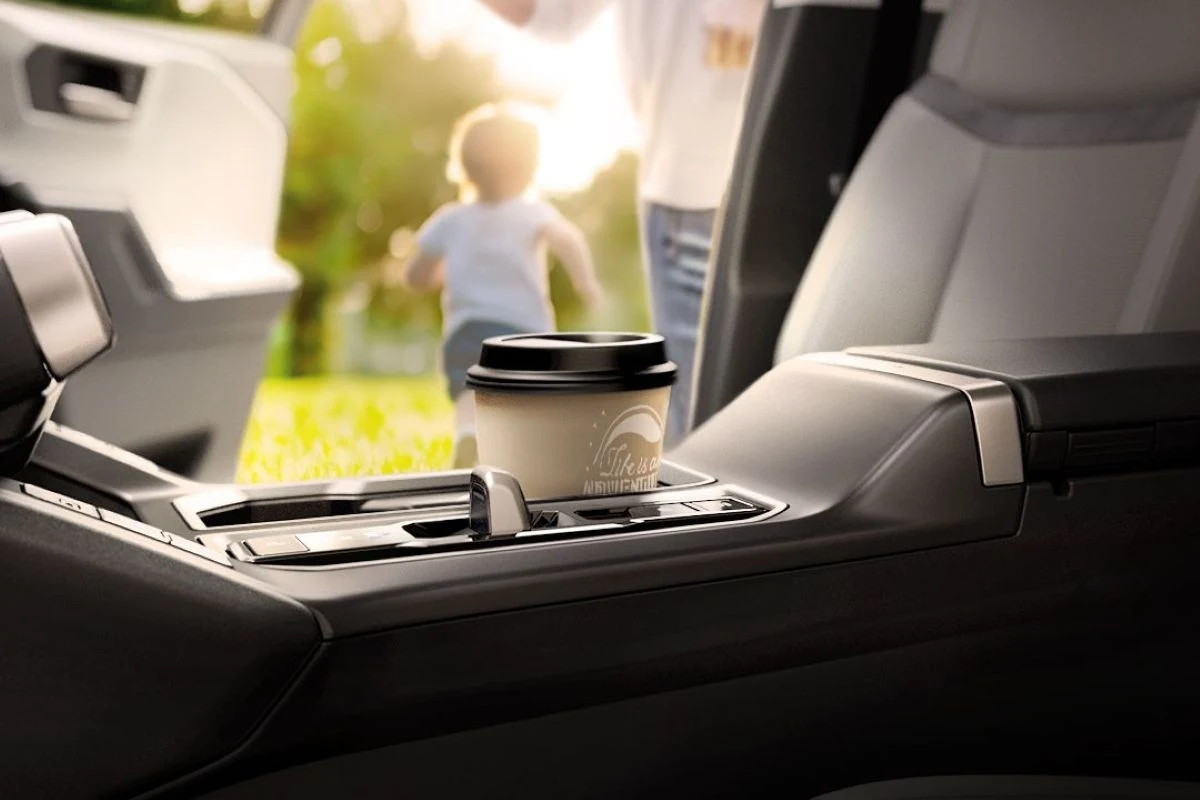 Kubek z kawą umieszczony w uchwycie w samochodowym podłokietniku. W tle rozmyty obraz rodzica z dzieckiem biegnącym po trawie, co sugeruje rodzinny moment. Wnętrze auta jest nowoczesne i eleganckie, z jasnym tapicerowaniem.