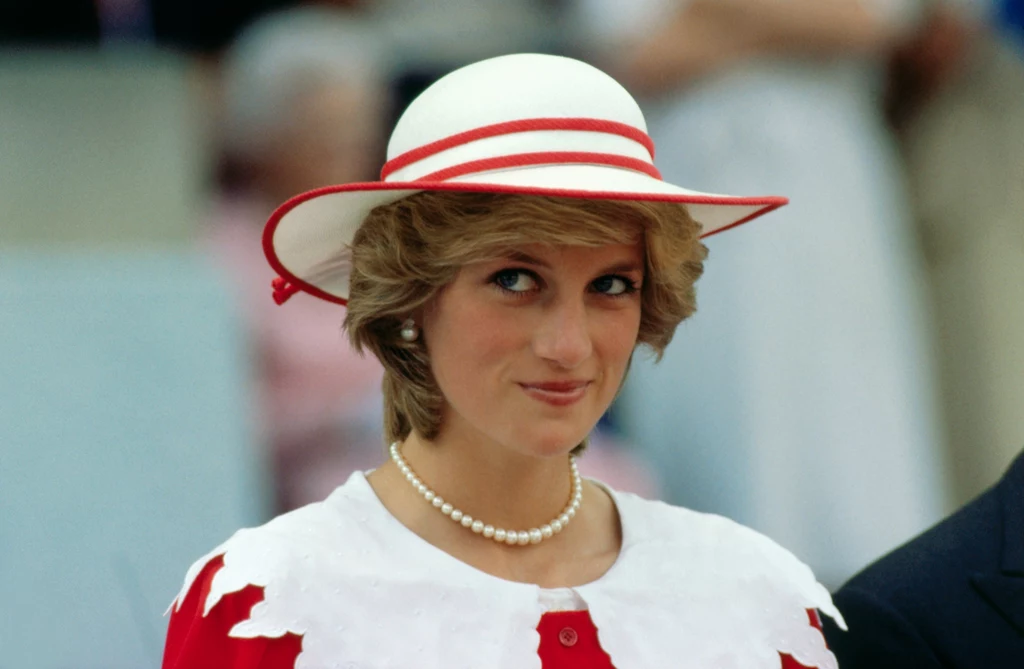 Księżna Diana w kapeluszu Kobieta w czerwono-białym stroju z perłowym naszyjnikiem i kapeluszem stojąca na zewnątrz. W tle nieostry obraz ludzi.